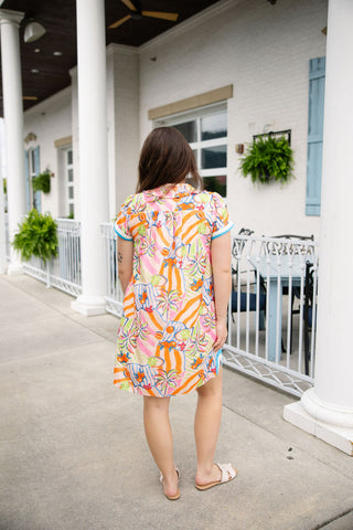 Back view of the Citrus Cabana Mini Dress, demonstrating the clean, continuous flow of the bright tropical pattern across the shoulders and relaxed shift frame. The mini-length curved hemline and the bright blue contrast bands on the cap sleeve cuffs are visible from behind. The model is turned away, showing her long straight brown hair and light beige slide sandals, emphasizing a breezy, effortless look perfect for summer travel.