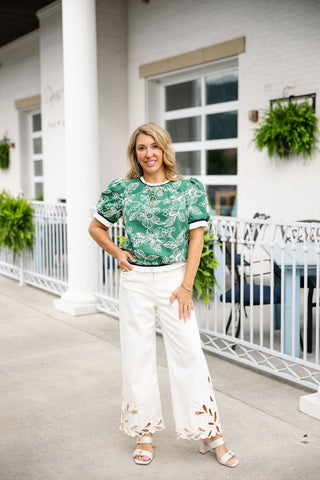 Woman posing in the Emerald Vine Blouse, a vibrant green woven top featuring detailed white botanical and floral embroidery. The blouse showcases short puff sleeves and a classic round neckline finished with a navy and white striped ribbed knit trim. The hem is also styled with the matching striped knit band and a delicate white fringe ruffle accent. Styled for a boutique resort look, tucked slightly into cream-colored wide-leg trousers featuring intricate laser-cut floral embroidery at the hem.

