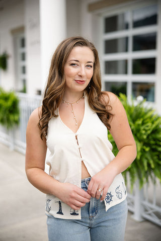An upper-body portrait view of a woman wearing the Harbor View Halter Top, highlighting the flattering deep V-neckline. The crisp off-white sleeveless top features a subtle split front with fabric loop button loops and a natural weave texture. Accessorized with a delicate gold drop pearl necklace, the maritime embroidery is visible along the split hemline at the natural waist.