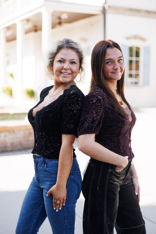 Two women posing back-to-back outdoors demonstrating color options for the short-sleeve paisley lace top. One model wears the classic black lace top with blue denim, and the other wears the dark plum purple lace top with black contrast-stitch corduroy pants.