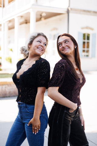 Two models standing back-to-back smiling, showing the side fit and sheer short sleeves of the paisley lace blouse in both classic black and deep plum purple colorways.