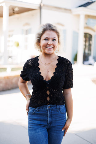 Upper body portrait of a woman wearing the black short-sleeve paisley lace top with scalloped edging and front keyhole cutouts, accessorized with gold hoop earrings and a delicate gold script necklace.