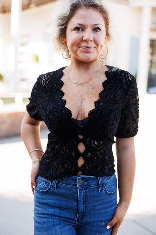 Woman standing outdoors wearing the short-sleeve paisley lace top in classic black. Features a deep scalloped V-neckline, puff sleeves, and diamond keyhole cutouts along the front button closure. Styled with high-waisted medium-wash blue jeans and gold hoop earrings.