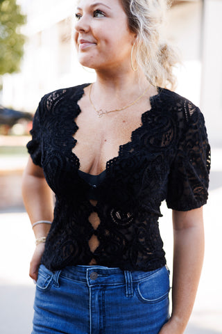 Upper body portrait of a woman wearing the black short-sleeve paisley lace top with scalloped edging and front keyhole cutouts, accessorized with gold hoop earrings and a delicate gold script necklace.