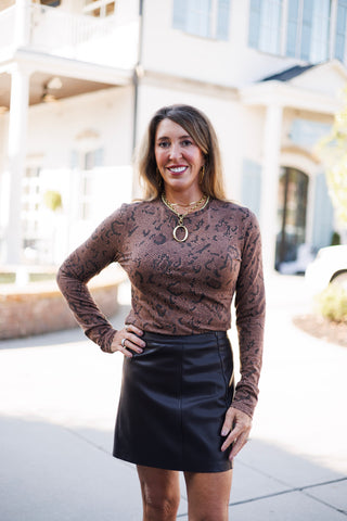 Detail shot focusing on the neckline and fabric texture of a brown long-sleeve snakeskin top paired with a brown leather skirt waist. Accentuated by layered gold chain necklaces with a prominent open-oval charm.