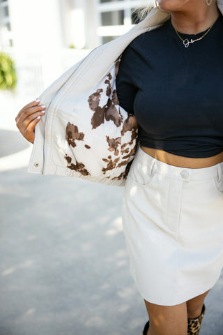 A close-up shot of the cream faux leather bomber jacket pulled open to reveal its bold brown and white cow-print inner lining. The view highlights the high-waisted cream faux leather mini skirt with front pockets and silver hardware details, paired with a fitted black top and gold chain necklace.