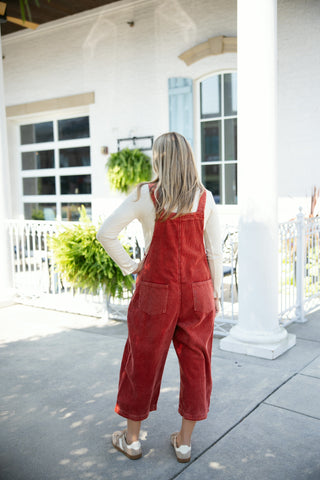 Back view of a woman wearing rust red wide-rib corduroy overalls, displaying the straight bib back construction and two oversized patch pockets across the seat. Styled with a long-sleeve cream top and beige lace-up sneakers on a sunlit concrete patio.
