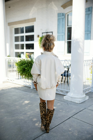 Back view of the cream faux leather two-piece set, displaying the slouchy, oversized bomber silhouette with elasticized waist detail and rolled cuffs exposing the cow-print lining. Paired with the matching mini skirt and styled with eye-catching knee-high leopard print boots on a patio walkway.