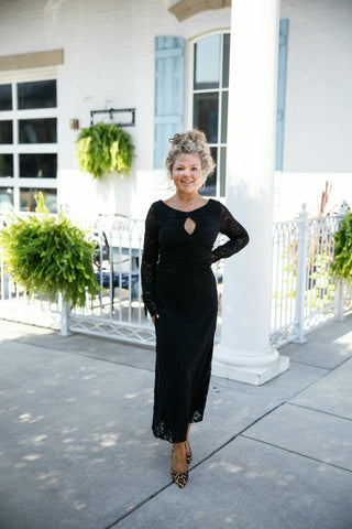 Woman posing outdoors in a black floral lace long-sleeve maxi dress featuring a keyhole cutout at the bust and a scoop neckline. The fitted column silhouette extends to an ankle-length scalloped hem, styled with point-toe leopard print pumps, gold hoop earrings, and a curly updo in front of a white building backdrop.