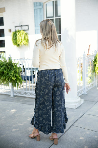 Back view of a woman wearing washed dark blue wide-leg denim crop pants featuring an all-over delicate cream floral print, ruffled side seams, and a ruffled raw-edge hemline. Paired with a fitted long-sleeve cream top and tan braided block-heel sandals on a sunny outdoor patio.