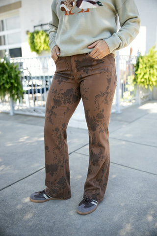 Mid-section close-up view highlighting high-waisted brown flare jeans featuring a subtle dark floral flocking pattern throughout. The pants feature classic five-pocket construction and a relaxed bootcut flare hem, styled casually with brown-and-white low-top sneakers and a sage green crewneck sweater.