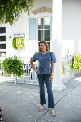 Full-length portrait of a woman posing outdoors in a washed blue ribbed corduroy top with puff sleeves and an embroidered pearl bow embellishment at the collar. She pairs the structured peplum top with matching dark blue raw-hem cropped flare jeans and metallic gold open-toe mules in front of a white building backdrop.