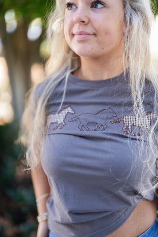 Close-up portrait shot showcasing the embroidered equestrian chest design of a charcoal grey crewneck t-shirt. The detail highlights three running wild horses with brown stitching and brown-and-white gingham patterned fill against the soft cotton fabric.