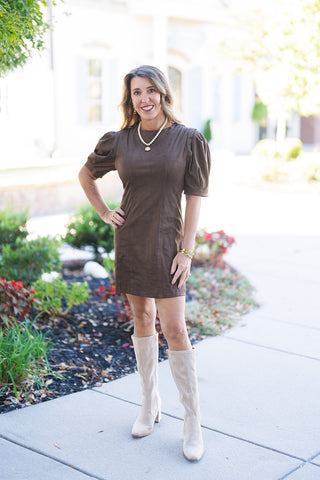 A woman posing outdoors on a concrete sidewalk in a chocolate brown faux suede mini dress featuring structured vertical panel stitching and short puff sleeves with banded cuffs. Styled with tall beige suede block-heel boots, a cream beaded statement necklace with a gold pendant, and stacked gold bracelets against a bright, softly blurred background.