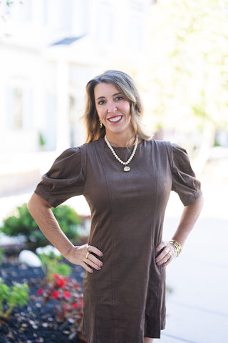 Three-quarter view of a smiling woman posing with hands on her hips, showcasing the tailored crew neckline and defined vertical seam detailing of the chocolate brown suede mini dress. Highlights the voluminous gathering at the shoulders of the elbow-length puff sleeves, accessorized with gold hoop earrings and a pearl-and-gold beaded necklace.