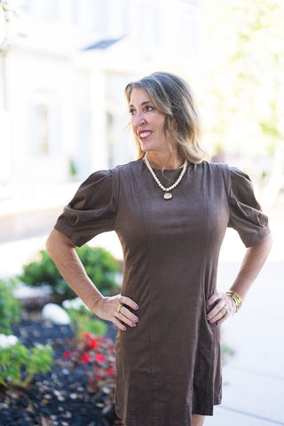 Medium portrait view highlighting the soft matte texture of the brown faux suede dress. The shot emphasizes the structured bodice seams, high crew neckline, and voluminous puff sleeves with tailored stitching details, accessorized with gold jewelry and framed by soft natural light outdoors.