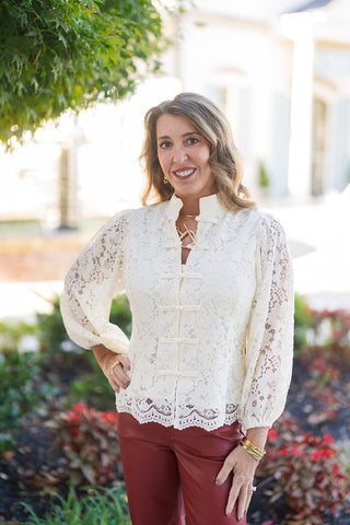 A waist-up shot showing the romantic floral lace pattern and tailored fit of the cream top. Features soft puff shoulders, blouson-style sleeves gather at elastic cuffed wrists, and a delicate scalloped eyelash lace trim finish along the bottom edge. Styled with deep red leather-look trousers and stack gold bracelets.