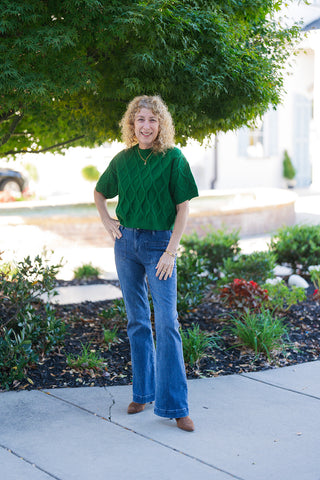 Woman posing outdoors on a sidewalk wearing a vibrant emerald green short-sleeve sweater featuring a textured diamond cable-knit pattern. Styled with high-waisted blue flare jeans with front patch pockets and brown pointed-toe suede boots under green leafy foliage.