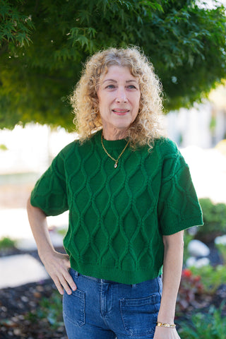 A portrait view of the short-sleeve knit sweater highlighting the ribbed crew neckline, dropped shoulder seams, and relaxed boxy silhouette. The rich emerald green top features raised lattice cable stitching, a ribbed hem, and wide ribbed sleeve cuffs, accessorized with a delicate gold pendant necklace.