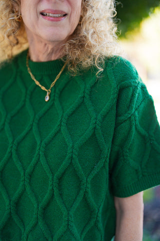 A close-up view emphasizing the intricate 3D diamond cable knit texture and soft yarn quality of the emerald green crewneck sweater. Captures the ribbed collar band and gold chain necklace with a small crystal stone charm.