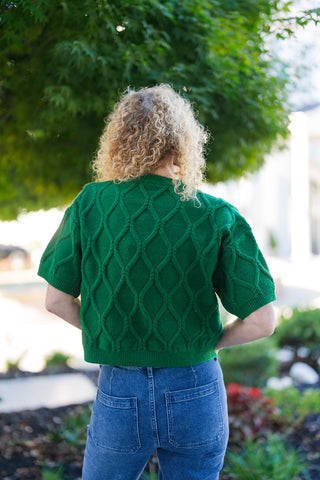 Back view of the short-sleeve green sweater showcasing the continuous diamond cable stitch pattern across the back and shoulders. Highlights the slightly cropped length resting neatly at the waistband of medium-wash blue jeans with back patch pockets.
