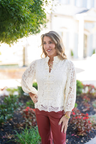 A portrait view of the cream lace blouse highlighting the mandarin collar with keyhole cutout detailing at the neck, gold statement jewelry, and traditional fabric knot front fasteners. The semi-sheer floral lace material is styled over a neutral base layer with structured puffy long sleeves.