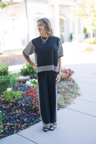A woman posing outdoors on a concrete sidewalk in a two-piece black knit lounge set featuring a short-sleeve top and matching wide-leg pants. The relaxed-fit top features vertical cream-and-black stripe panels down the sides and short sleeves, balanced by a horizontal striped border along the hem. The trousers display a vertical side stripe and horizontal striped cuffs, complete with black strappy sandals and a chunky gold toggle necklace.