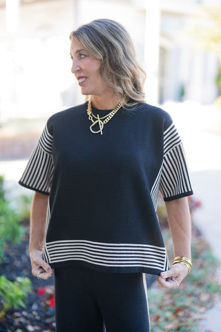 Medium shot of a woman modeling the black short-sleeve knit top, highlighting the dropped-shoulder construction and crew neckline. The fine-gauge knit fabric is detailed with contrast cream vertical stripes along the sleeves and side seams, as well as a multi-stripe horizontal hem border. Styled with stacked gold bangles and a statement gold chain necklace.