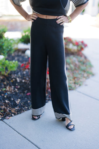 Front view focused on wide-leg black knit trousers styled with a matching short-sleeve knit top. The pants feature a comfortable high-waisted elastic band, slender cream side stripes running down each leg, and horizontal cream-and-black striped border trim along the wide hemline. Paired with black open-toe strappy sandals on an outdoor walkway.