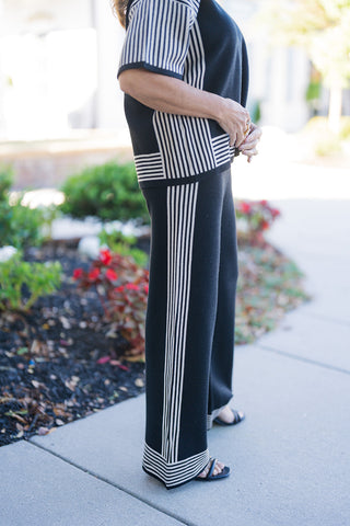 Side profile shot highlighting the geometric stripe detail of a black and cream fine-knit two-piece set. Showcases the continuous cream-and-black vertical side stripe extending down the wide-leg pants and matching short sleeve top, accented by horizontal striped hem borders. The model wears black strappy heel sandals on a paved sidewalk setting.