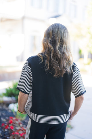 Back view of the black short-sleeve knit top showing the solid black back panel flanked by vertically striped sleeves. The horizontal cream-and-black striped hem band provides a structured finish over matching knit pants, set against a soft-focus outdoor walkway background.