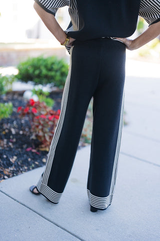Back view of a woman posing with hands on hips, showcasing high-waisted black wide-leg knit pants with vertical cream side stripes. The design features clean back styling with horizontal striped cuffs at the wide hem. Styled with a matching loose-fit knit top and black open-toe shoes on a sunny outdoor pathway.