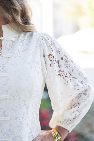 A detailed close-up shot emphasizing the intricate floral lace embroidery, soft shoulder gather seam, and fabric frog button closure detailing down the center. Displays the delicate texture of the sheer lace puff sleeve gather and elasticated cuff.