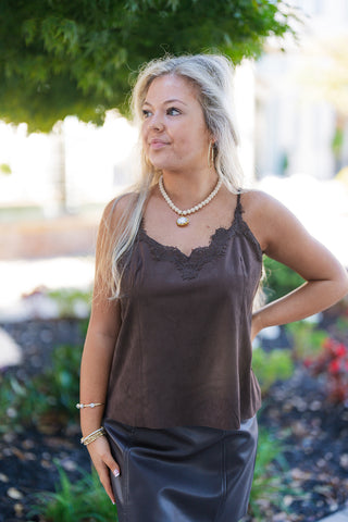Three-quarter view of a woman modeling the dark brown faux suede Cami top, highlighting the scalloped lace detailing trim around the V-neckline. The top features a relaxed silhouette and slender shoulder straps, styled tucked over a high-waisted dark leather mini skirt with gold hoop earrings and a beaded stone necklace.