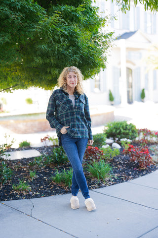 Woman posing outdoors standing on a concrete path wearing an oversized forest green and navy blue plaid button-down shacket. Styled over a beige base layer top, classic dark-wash slim-fit jeans, and cozy beige shearling-lined mule slippers. She stands in front of a lush green tree and landscaped garden bed with her hand casually resting in her pocket.