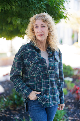 A portrait view highlighting the oversized plaid shirt-jacket featuring a classic collar, brown buttons, and two front chest flap pockets with diagonal bias-cut plaid details. Made from a textured green, navy, and cream tartan plaid pattern, layered over a neutral crewneck shirt. The woman with blonde curly hair has one hand tucked into her denim jeans pocket.