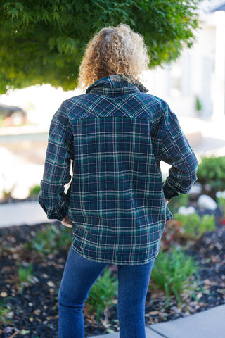 Back view of the oversized plaid shacket showcasing the continuous forest green and navy plaid design across the yoke and back panel. Features dropped shoulders, a relaxed loose-fit silhouette, long sleeves, and a curved hemline covering the upper hips. Paired with dark blue fitted jeans outdoors under tree foliage.