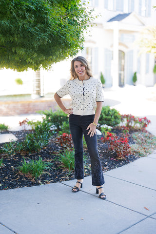Woman posing outdoors in an outfit featuring black cropped flare denim jeans with intricate tonal embroidery and subtle stud details along the hem, paired with a short-sleeve cream cardigan with black polka dots and black strappy heeled sandals.