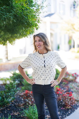 A waist-up shot highlighting the short-sleeve button-down cardigan top with small black polka dots on a cream knit body. Features slight puff shoulders, a ribbed crew neckline with dark contrast trim, matching ribbed hem and cuffs, and small white front buttons. Styled with stack bracelets and gold jewelry.