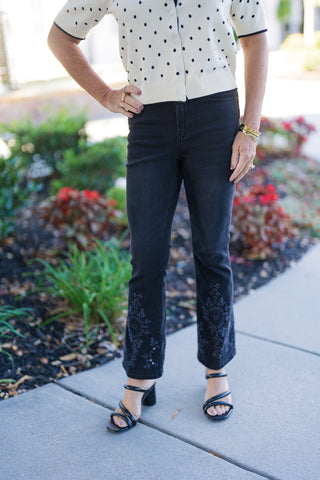 Close-up front view of the black cropped flare denim jeans and cream polka-dot short-sleeve cardigan, highlighting the tailored fit, waistline, and detailed embroidered hem design.