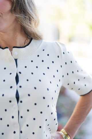 A detailed close-up shot showcasing the fine knit texture, black polka dot print, and delicate gather at the shoulder seams. Highlights the dark contrast tipping on the crew neck and sleeve edges, white front closure buttons, and fine ribbed trim along the collar.