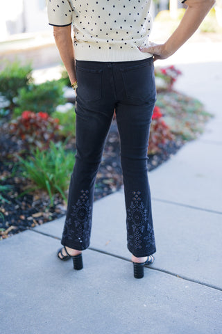 Back view of the outfit, showcasing the clean silhouette of the black denim jeans with back pockets and the embroidered hem details extending down to the ankles, worn with a cream polka-dot cardigan and black heeled sandals.