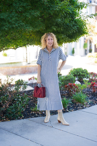 Woman posing outdoors on a sidewalk wearing a relaxed-fit blue and white gingham check midi shirtdress. Designed with short cuffed sleeves, an open split neckline with a flat notch collar, side hem slits, and a straight silhouette. Styled with cream Western-style pointed boots, a deep red velvet handbag, and gold jewelry in front of lush garden foliage.
