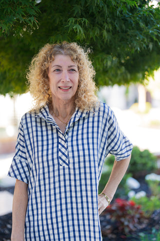 A waist-up view of the blue and white gingham shirt dress highlighting the open popover-style split neck placket with a diagonal bias-cut pattern detail. Shows the relaxed drop-shoulder cut, cuffed short sleeves, and soft seersucker-like woven pattern texture, accessorized with a delicate gold chain necklace.