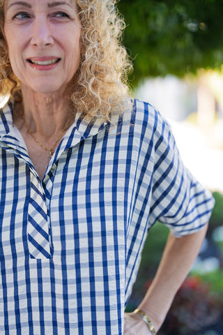 A close-up portrait shot emphasizing the woven detail of the royal blue and white gingham check pattern fabric. Highlights the folded notch collar design, bias-cut placket seam, and short sleeves styled on a woman with blonde curly hair.