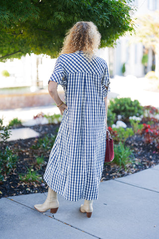 Back view of the blue and white gingham midi dress showcasing a diagonal bias-cut back yoke and a center back box pleat for an easy, flowing drape. The dress flows down to a midi length, styled outdoors with light beige snake-print Western boots and a maroon handbag.