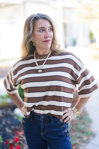 Three-quarter front view of a woman with her hands on her hips, showcasing a brown and cream striped knit top with dropped shoulder half-sleeves and a crew neckline. She is styled with layered gold and beaded necklaces, gold hoop earrings, and stacked bracelets. Her high-rise dark-wash denim jeans feature decorative metallic buttons along the front pockets, capturing a casual, accessorized look on an outdoor pathway.