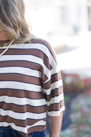 Close-up cropped shot highlighting the texture of the striped knit top, showing the fine-gauge knit pattern with alternating brown and cream horizontal stripes. Focuses on the dropped shoulder seam, relaxed half-sleeve, and ribbed crew neckline. The subject wears layered necklaces, including a beaded strand, with the dark indigo denim of the high-rise jeans subtly visible at the bottom against a soft, out-of-focus outdoor backdrop.