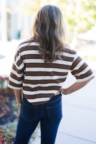 Back view of a woman posing outdoors in a brown and cream striped knit top paired with dark-wash jeans, highlighting the casual, relaxed fit. The short-sleeve top features horizontal stripes and a loose, boxy cut that sits natural at the waist. Fitted dark-wash denim jeans are visible with back pockets and contrast stitching, set against a blurred outdoor walkway with natural daylight.