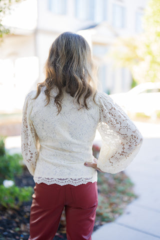 Back view of the ivory lace blouse displaying the continuous sheer lace floral overlay across the shoulders and back panel. Highlights the gentle gather at the top of the balloon sleeves and the semi-sheer scalloped lace hemline resting neatly over dark red leather pants.